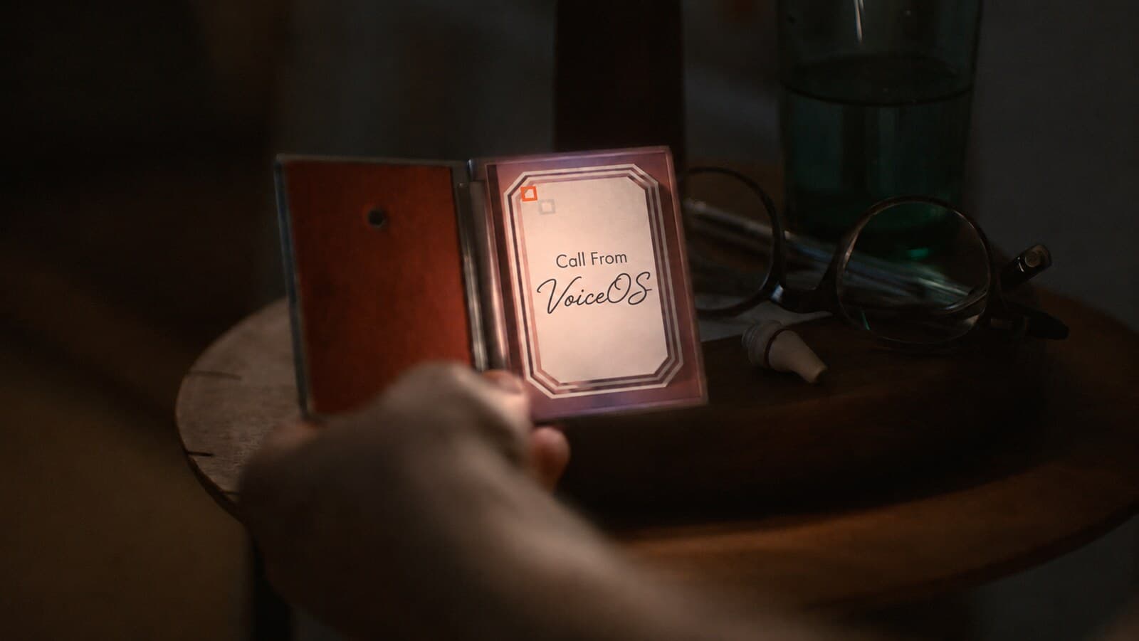 A Her-style folding handheld device on a nightstand showing an incoming call from VoiceOS
